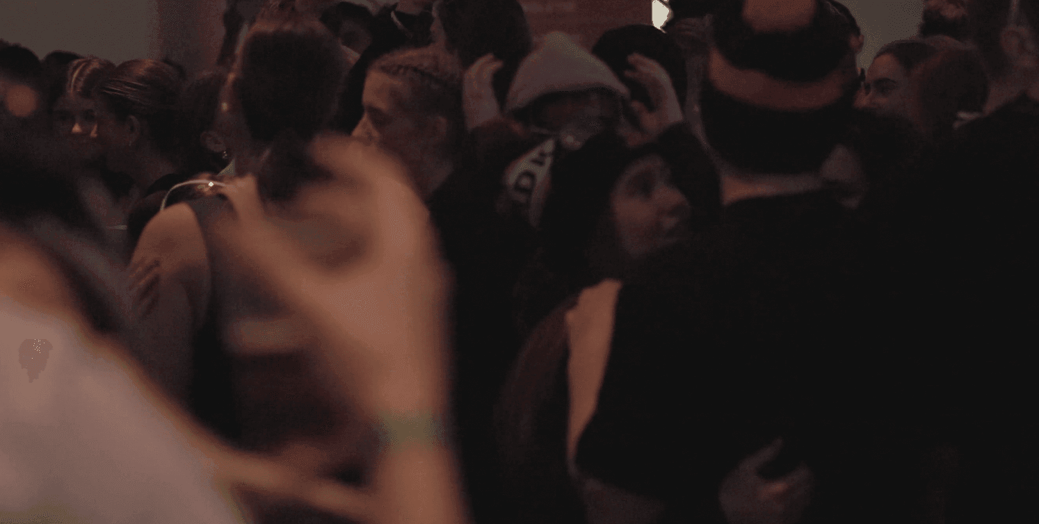 People closely gathering in a dimly lit space, some wearing hats and holding drinks.