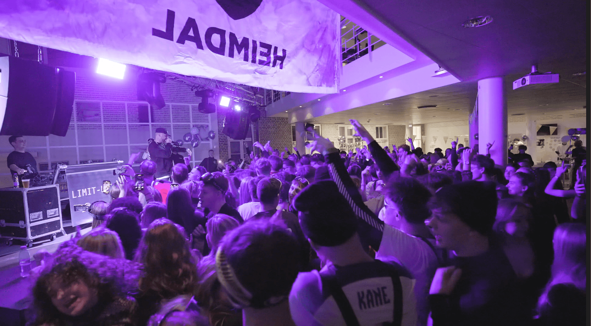 Crowd enjoying a live music performance indoors, with purple stage lighting and a banner overhead.